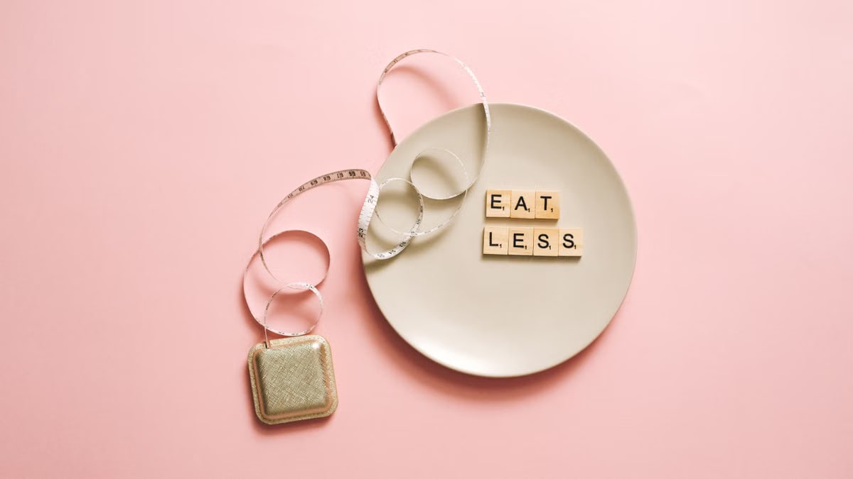 A motivational image featuring a plate with scrabble tiles spelling 'Eat Less' on a pink background.